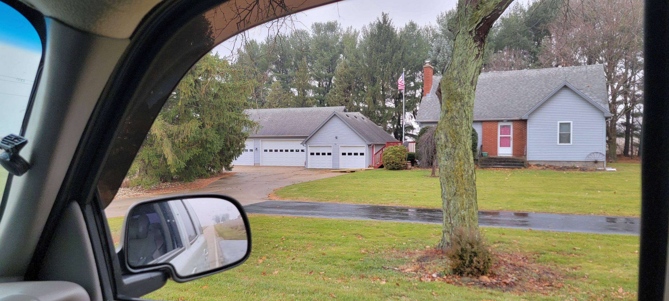 View of a suburban house and garage with lush greenery, seen from a car window on a rainy day.